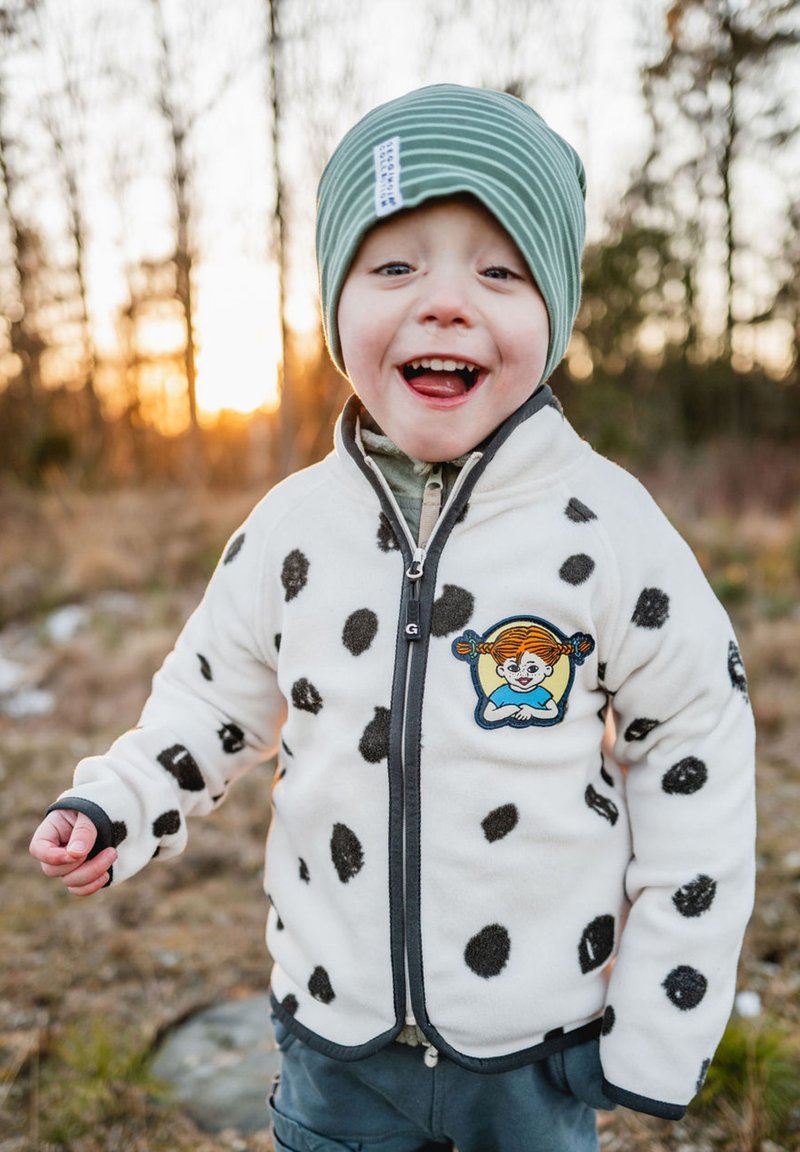 Fleece-takki, jossa on mustat pilkut valkoisella, vetoketjulla edessä ja harmaalla reunuksella. Sisältää kirjotun patchin hahmosta. Vihreä raidallinen pipot.