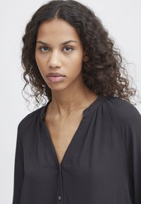 Black button-up blouse with a V-neckline, soft texture, and subtle pleats on the shoulders. Features small buttons and a fluid drape.