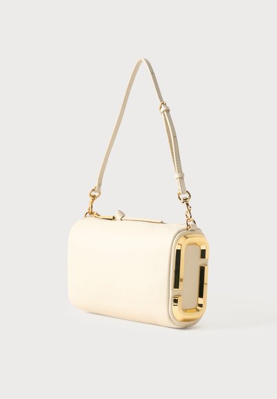 Κρεμ δερμάτινη τσάντα ώμου με ρυθμιζόμενο λουρί και μεγάλο λογότυπο από χρυσό μέταλλο στο πλάι, μπροστά σε ουδέτερο φόντο.