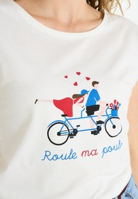 Camiseta blanca con un gráfico de una bicicleta tándem azul, con una mujer en un vestido rojo y un hombre en una camisa azul. El texto dice "Roule ma poule."