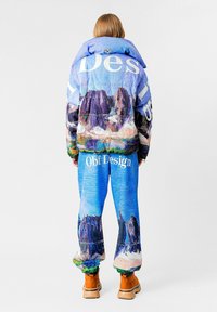 Chaqueta y pantalones acolchados con un estampado de paisaje montañoso en azul y varios colores, complementados con un texto audaz. Botines marrones completan el conjunto.