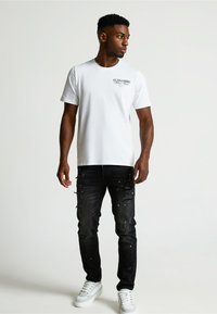 Biele tričko s krátkym rukávom a logom vpredu, v kombinácii s čiernymi roztrhnutými džínami s rozstrapkanými okrajmi a bielymi akcentmi, a bielymi teniskami.