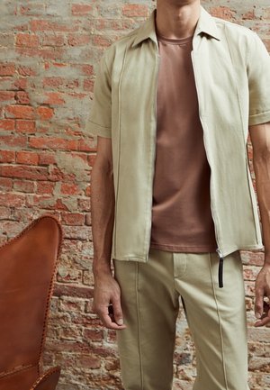 Beżowa kurtka na zamek błyskawiczny, krótki rękawek, noszona na brązowym t-shircie, w zestawie z dopasowanymi beżowymi spodniami. Gładki materiał, prosty fason, minimalne akcenty.