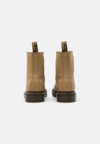 Un paio di stivaletti in pelle color beige con suole nere e cuciture gialle, visti da dietro su uno sfondo bianco.