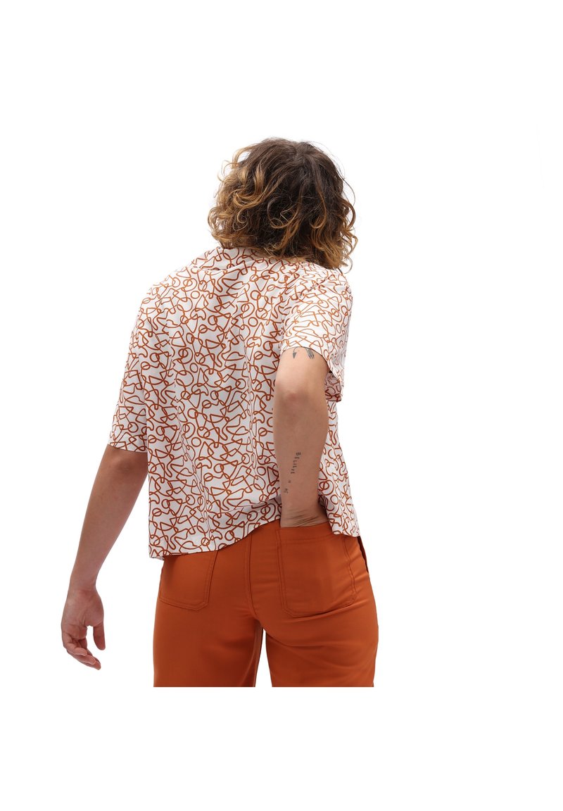 Personne aux cheveux bouclés portant une chemise blanche avec des lignes abstraites marron et un pantalon orange, vue de dos avec une main dans la poche.