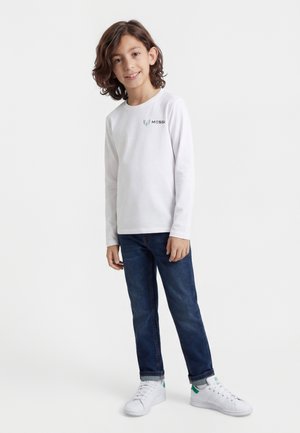 Dreng med skulderlangt hår iført hvid langærmet skjorte med "Messi"-logo, blå jeans og hvide sneakers mod en hvid baggrund.