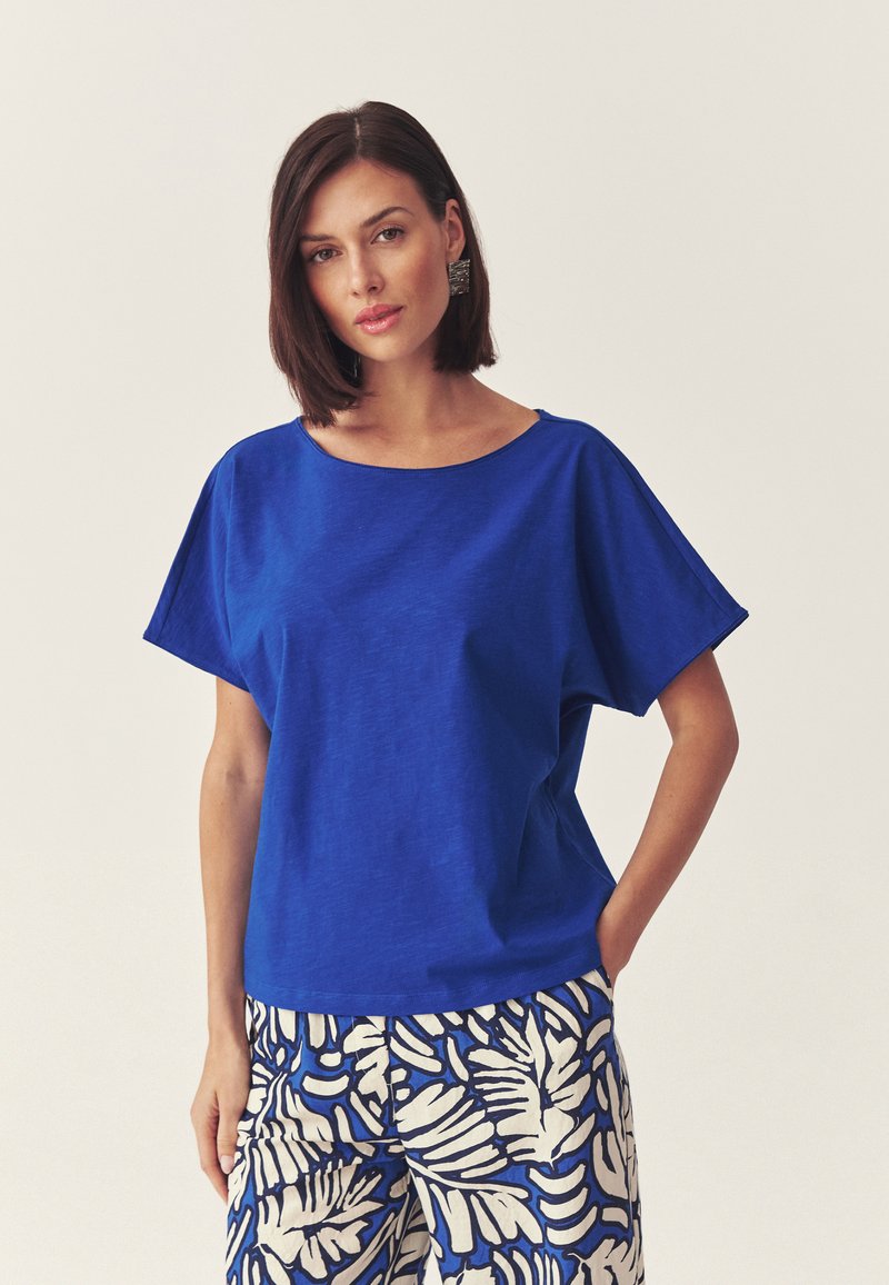 Femme aux cheveux bruns courts portant un haut bleu ample et un pantalon blanc à motifs de feuilles bleues, debout devant un fond clair uni.