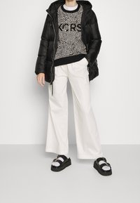 Persona vestida con una chaqueta acolchada negra, suéter con estampado de animales, pantalones amplios blancos y sandalias de plataforma negras con calcetines blancos, de pie contra un fondo simple.