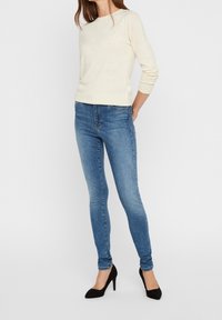 Blusa de manga larga cremosa combinada con jeans ajustados azules. Tejido texturizado y diseño entallado. Zapatos de tacón negro completan el look.