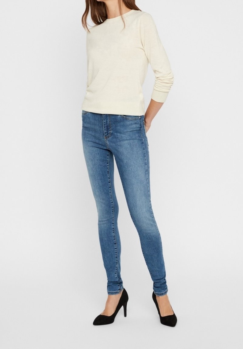Blusa de manga larga cremosa combinada con jeans ajustados azules. Tejido texturizado y diseño entallado. Zapatos de tacón negro completan el look.