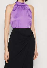 Top halter in satin viola con collo alto, abbinato a una gonna nera con accentuazione a nodo attorcigliato. Texture liscia e silhouette aderente.