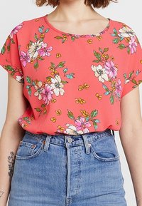 Korallefarbene Bluse mit lässiger Passform, kurzen Ärmeln und Rundhalsausschnitt. Mit floralen Mustern in Pink, Weiß und Grün auf einem glatten Stoff.