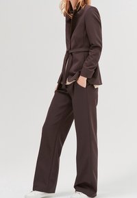 Kobieta ubrana w ciemnobrązową marynarkę z paskiem, dopasowane szerokie spodnie oraz białe trampki, stojąca z jedną ręką w kieszeni.