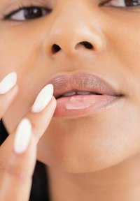 Nahaufnahme von Lippen, die mit einem glänzenden nude Lippenstift geschminkt sind. Kurze, ovale weiße Nägel berühren die Lippen, und die Haut hat eine glatte, gleichmäßige Textur.