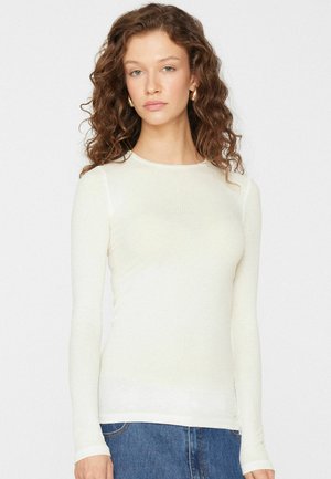 Žena s kudrnatými vlasy, která nosí zlaté kruhové náušnice, přiléhavé bílé tričko s dlouhým rukávem a modré džíny, stojící před jednobarevným pozadím.