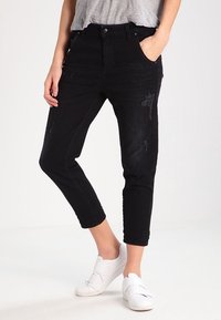 Schwarze Denim-Jeans mit einer taillierten Passform, die mit Used-Effekten und einer verkürzten Länge versehen sind. Getragen mit einem grauen Oberteil und weißen Sneakers.