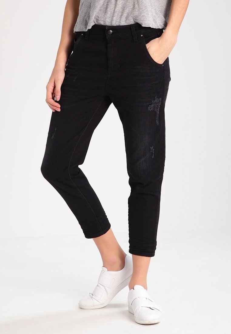 Schwarze Denim-Jeans mit einer taillierten Passform, die mit Used-Effekten und einer verkürzten Länge versehen sind. Getragen mit einem grauen Oberteil und weißen Sneakers.