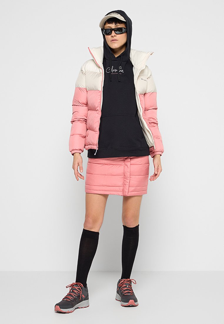 Person in einer rosa und weißen Steppjacke und einem Rock, schwarzem Hoodie, schwarzen knielangen Socken, grau-rosa Sneakers, Sonnenbrille und beiger Kappe.