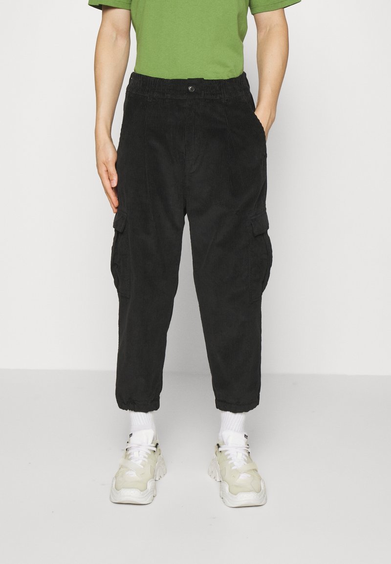 Personne portant un pantalon cargo en velours côtelé noir, des baskets blanches, des chaussettes blanches et un t-shirt vert à manches courtes, debout devant un fond blanc.