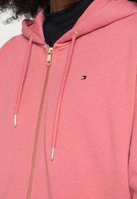 Detailní záběr osoby v růžové mikině na zip se zlatým zipem a malým červeným, bílým a modrým logem na hrudi přes černé tričko.