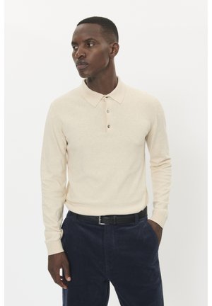 Beige Langarm-Polohemd mit Kragen, Knopfleiste und subtiler Textur, kombiniert mit dunkelblauen Cordhosen und einem schwarzen Gürtel.