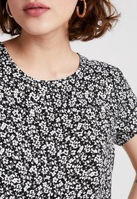 Černobílé tričko s květinovým vzorem, kulatým výstřihem a krátkými rukávy. Látka působí lehce a má hladkou texturu.