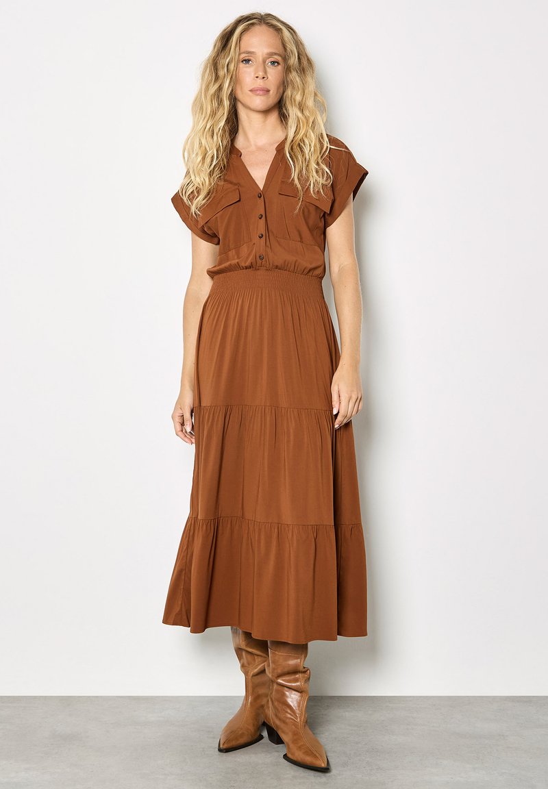 Robe longue marron à volants avec manches courtes, devant à boutons, taille élastique et tissu lisse. Associée à des bottes hautes marron clair jusqu'aux genoux.