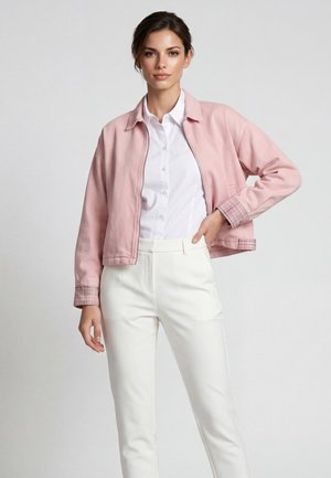 Frau trägt eine hellrosa Jacke über einem weißen Hemd mit Knöpfen und weißen Hosen, steht mit einer Hand in der Hüfte vor einem einfarbigen Hintergrund.
