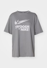Šedé bavlněné tričko s kulatým výstřihem, krátkými rukávy a velkým bílým logem "Swoosh" a textem "SWOOSH BY NIKE" potištěným na přední straně.