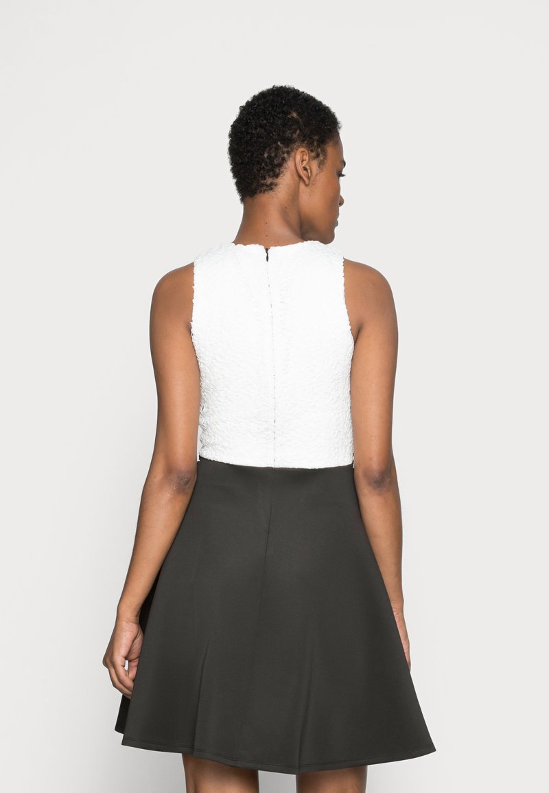 Weißes strukturiertes Oberteil mit hohem Kragen und einem schwarzen ausladenden Rock. Verfügt über einen Reißverschluss auf der Rückseite und ist ärmellos.