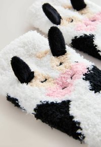 Fuzzy, vita och svarta tofflor med struktur utformade som en ko, med svarta öron, rosa nos och broderade ansiktsdetaljer.