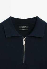 Marinblå bomullspolo med dragkedja, ribbad textur och klassisk krage. Etiketten anger 100% bomullsmaterial.