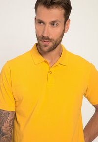 Žluté polo tričko se strukturovaným límečkem, krátkými rukávy a jedním knoflíkem. Hladká bavlněná tkanina s čistým, jednobarevným designem.