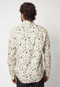 Uomo con capelli ricci che indossa una camicia crema decorata con ghirlande di Natale, alberi, bastoncini di zucchero, regali, vischio e agrifoglio visto da dietro.