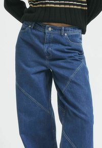 Tumesinised denim püksid vabalt lõikega, diagonaalsete õmbluste detailidega ja esikottidega. Mudel kannab musta triibulist sviitrit.