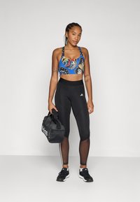 Αθλητικό σουτιέν με μπλε, πολύχρωμο σχέδιο πεταλούδας, σε συνδυασμό με μαύρα κολάν που διαθέτουν δικτυωτά πάνελ και τρεις λευκές ρίγες. Μαύρη τσάντα γυμναστηρίου.