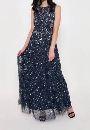 Granatowa wieczorowa sukienka bez rękawów z misternym wzorem z srebrnych cekinów, długa tiulowa spódnica sięgająca podłogi, noszona z czarnymi sandałami na otwartym palcu.