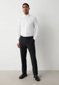 Weiß gestreiftes Hemd mit spitzem Kragen, kombiniert mit schwarzen maßgeschneiderten Hosen und schwarzen Lederschuhen, auf einem neutralen Hintergrund stehend.