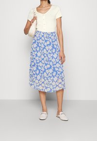 Ljusgul figurpassad kortärmad topp med knappar, i kombination med en flödande blå blommig midi-jurkd. Vita slip-on skor och en liten väska.