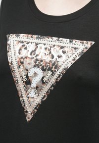 Črna majica s kratkimi rokavi s veliko trikotno grafiko v leopardjem vzorcu in srebrnimi okrasnimi žeblji. Vključuje napise "GUESS®," "WASHED," "JEANS" ter vprašaj.