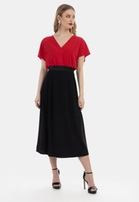 Top rosso a maniche corte con scollatura a V, abbinato a una lunga gonna nera con una vita strutturata. La modella indossa sandali neri con tacco.