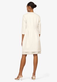 White knee-length dress with 3/4 sleeves, textured fabric, flared skirt, and frayed hem, paired with white strappy heels.