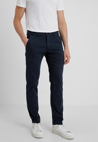 Pantalones de algodón azul marino con un corte regular, que cuentan con bolsillos laterales, cremallera y trabillas para cinturón, combinados con zapatillas blancas.