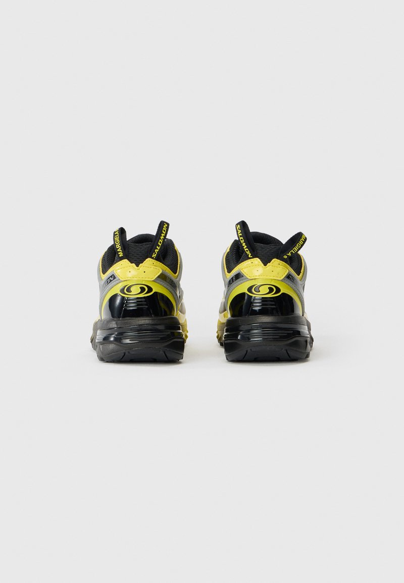 Par af sorte og gule Salomon-sneakers vist fra baggrunden, der viser mærke-trækstropper og tykke polstrede såler på en ensfarvet baggrund.