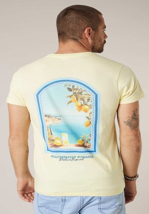 Mann trägt ein hellgelbes T-Shirt mit einem Rückenaufdruck einer mediterranen Meeresküstenansicht, Zitronen und dem Text "Mediterranean Sundance Deluxe Original".