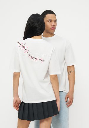 To personer iført hvide t-shirts, den ene set forfra med synligt tatoveret arm, den anden set bagfra med design af lyserøde kirsebærblomster og tekst.