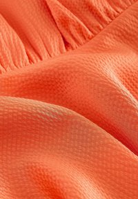 Gros plan d’un tissu texturé corail-orange avec un détail élastique froncé créant des plis doux et un motif en relief rappelant une gaufre.