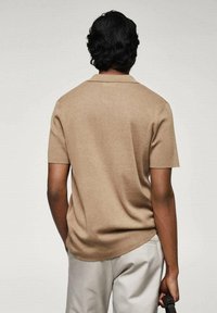 Homme aux cheveux bouclés foncés portant une chemise beige à manches courtes et un pantalon clair, se tenant de dos, tenant une sangle noire dans sa main droite.