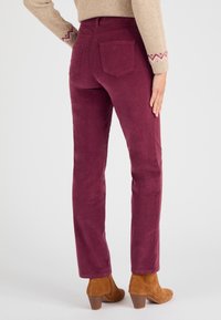 Bordeaux corduroy broek met een aansluitend ontwerp, voorzien van twee achterzakken en gedragen met bruine haklaarzen. Textuur van het materiaal is duidelijk zichtbaar.