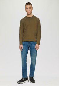 Camisa de manga larga verde oliva con textura acanalada, combinada con jeans de mezclilla azul claro y zapatillas deportivas negras con acentos blancos.